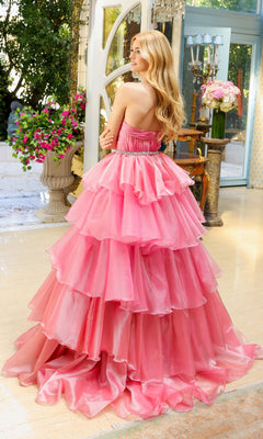 A woman with long blonde hair stands indoors, facing away amid flowers and elegant decor, wearing the Long Prom Dress 29546 by Ava Presley—a strapless pink ball gown with a sweetheart neckline and tiered skirt.