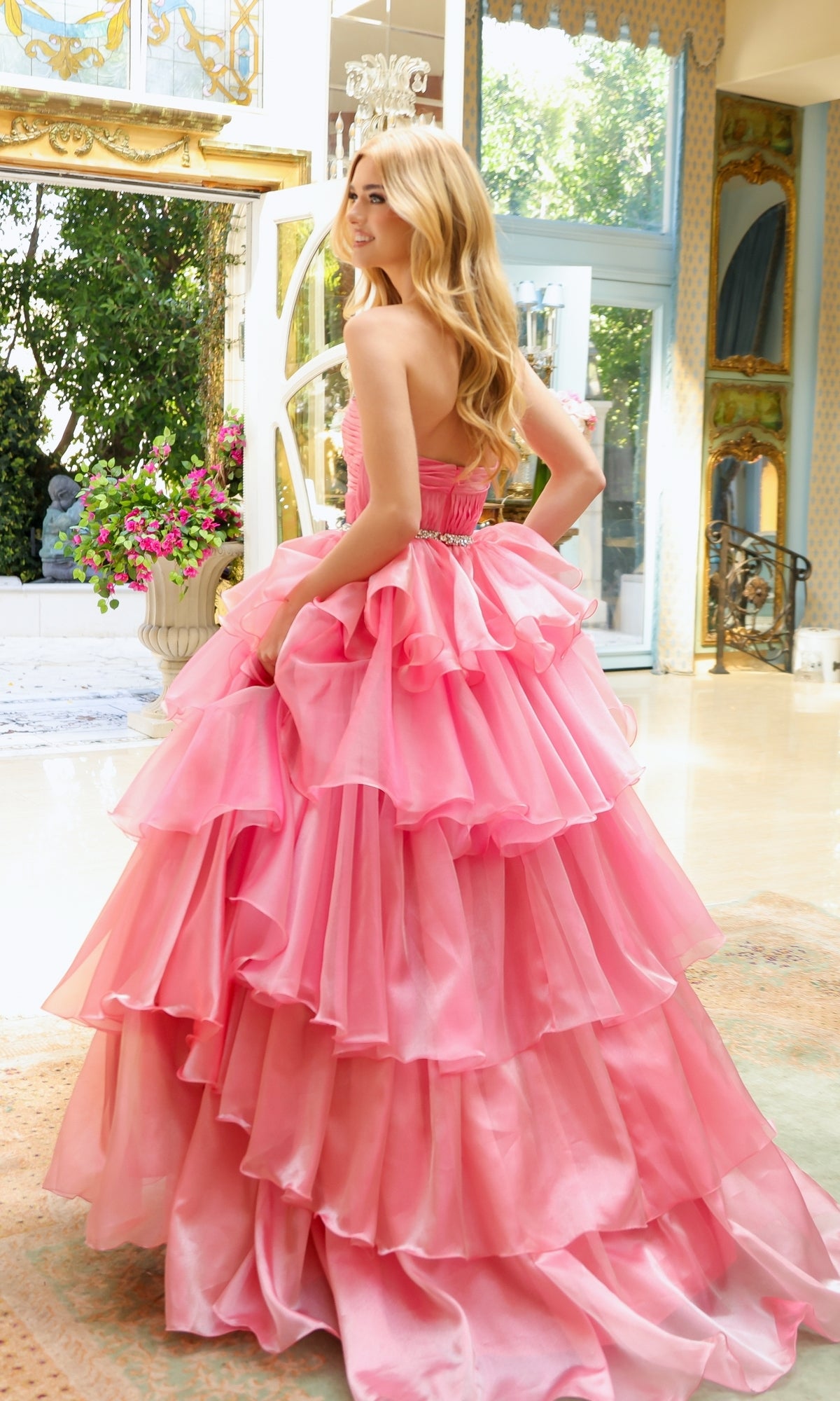 A woman with long blonde hair wears the Ava Presley Long Prom Dress 29546, a strapless sweetheart layered ruffle ball gown, standing indoors by large windows with sunlight and greenery outside.