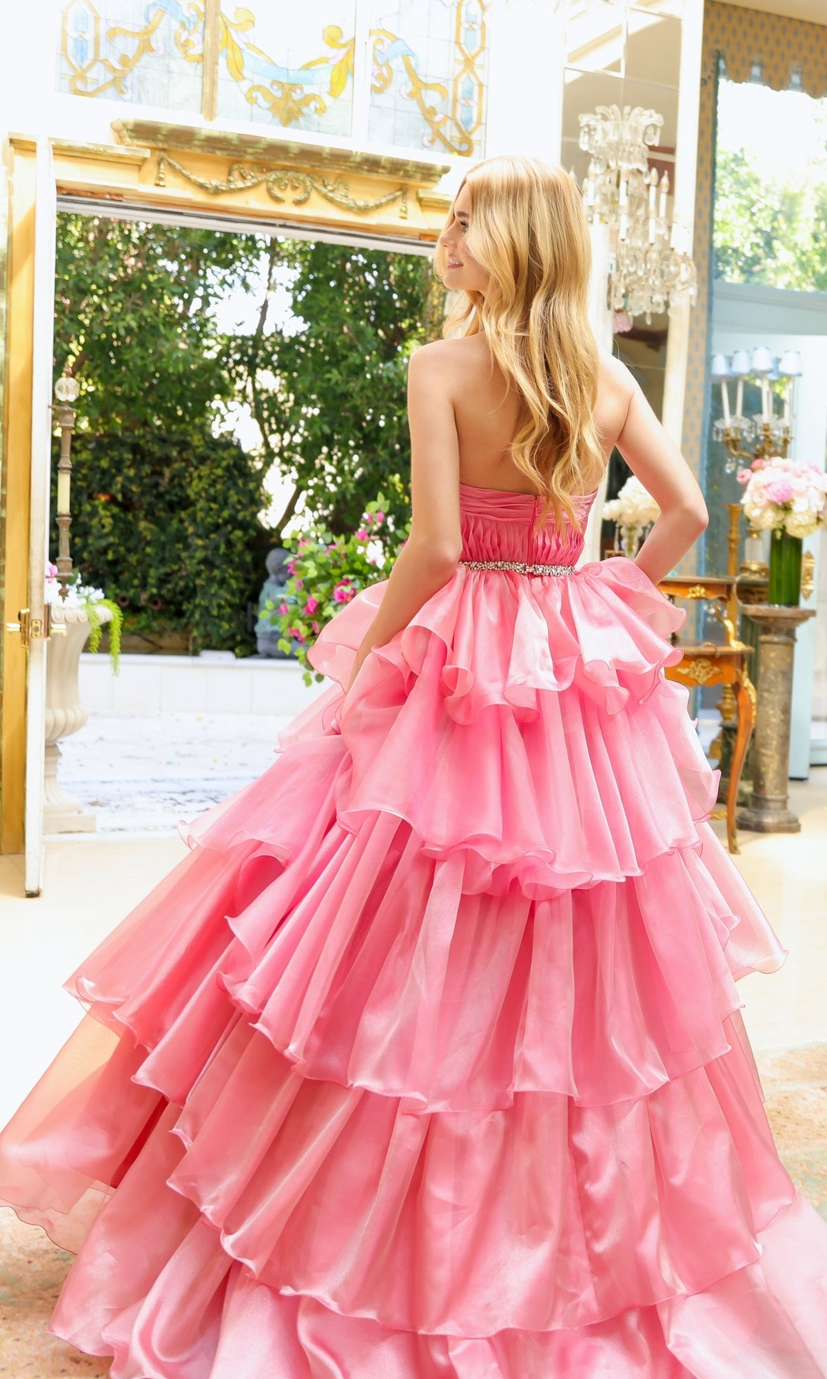 A woman wearing the Long Prom Dress 29546 by Ava Presley, featuring a strapless sweetheart neckline and layered ruffle ball gown, stands indoors facing away as sunlight streams through large windows.