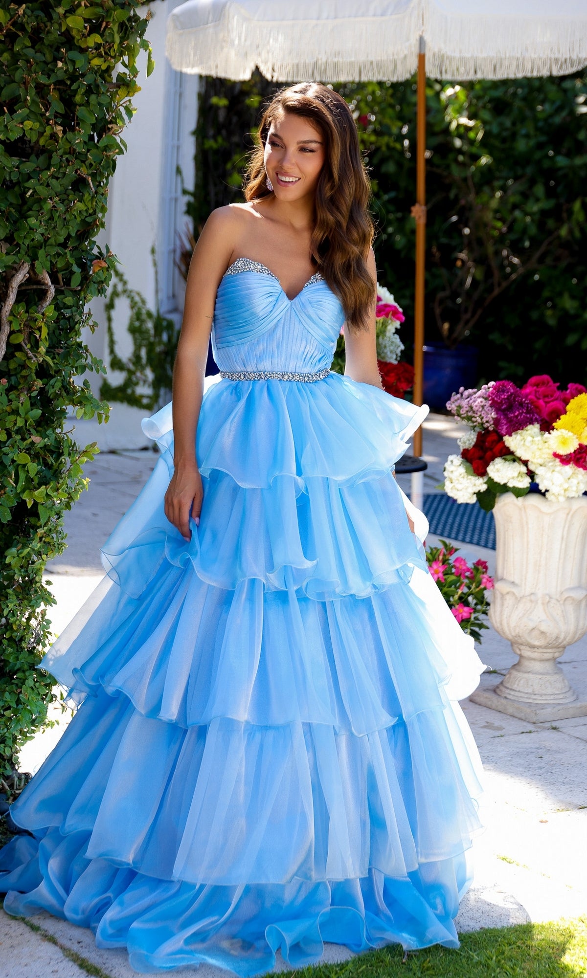 A woman stands outdoors next to colorful flowers under a white umbrella, wearing the Long Prom Dress 29546 by Ava Presley—a strapless blue ball gown with a sweetheart neckline and jeweled waistband.
