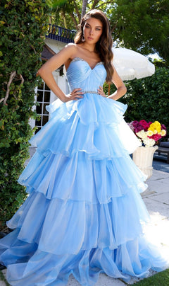 A woman stands outdoors wearing the Long Prom Dress 29546 by Ava Presley, a strapless sweetheart ball gown with layered ruffles and a jeweled waistband. Trees, a white umbrella, and a flower arrangement are in the background.