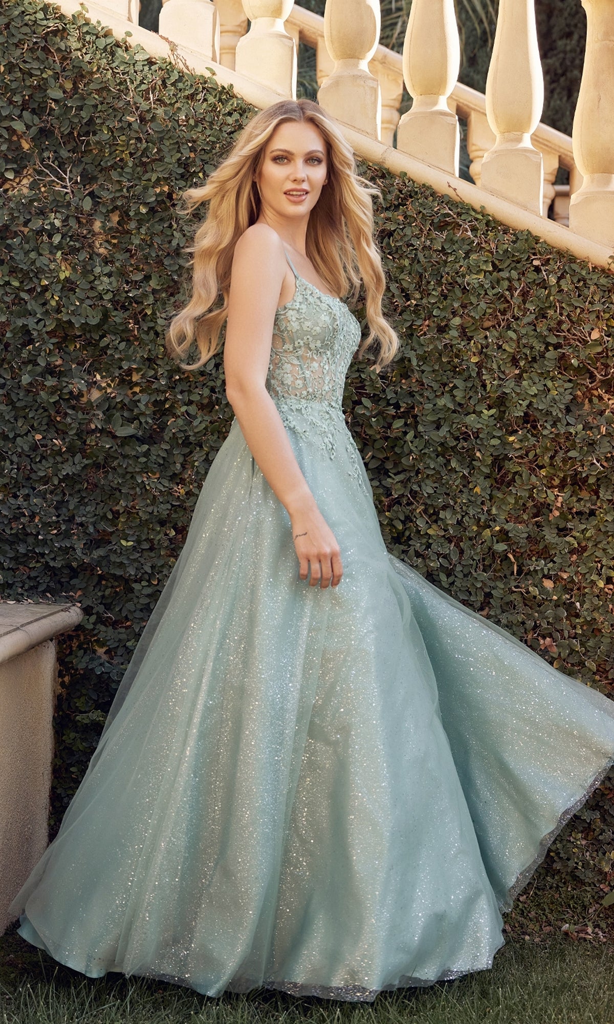 A woman in the Juliet 295 Lace-Up Long A-Line Prom Dress stands outdoors by a staircase and green foliage, facing the camera.