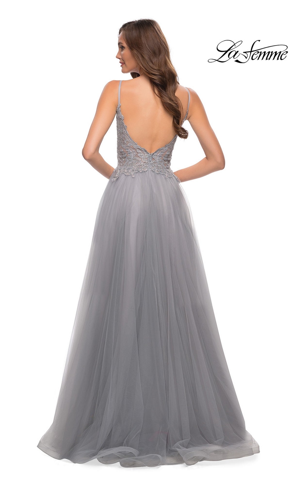 A woman stands with her back to the camera in a light gray La Femme Lace-Bodice Long A-Line Prom Dress 29686, featuring an embroidered lace bodice, deep V-back, and flowing tulle skirt.
