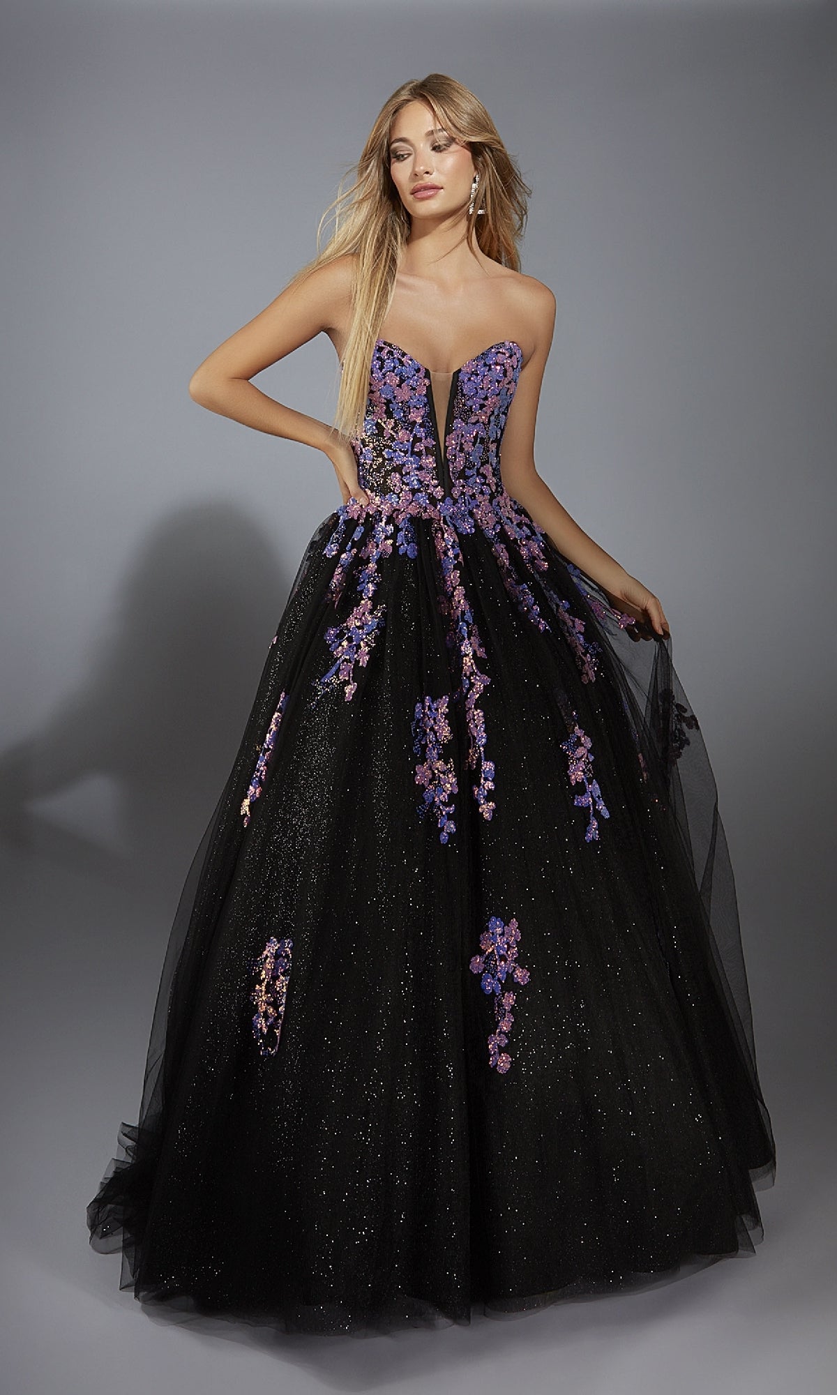 A woman wears the Strapless Glitter Prom Ball Gown: Aerie Couture 297, featuring a strapless black floral sequin design with purple and pink appliqués and a deep V neckline, standing against a gray background.