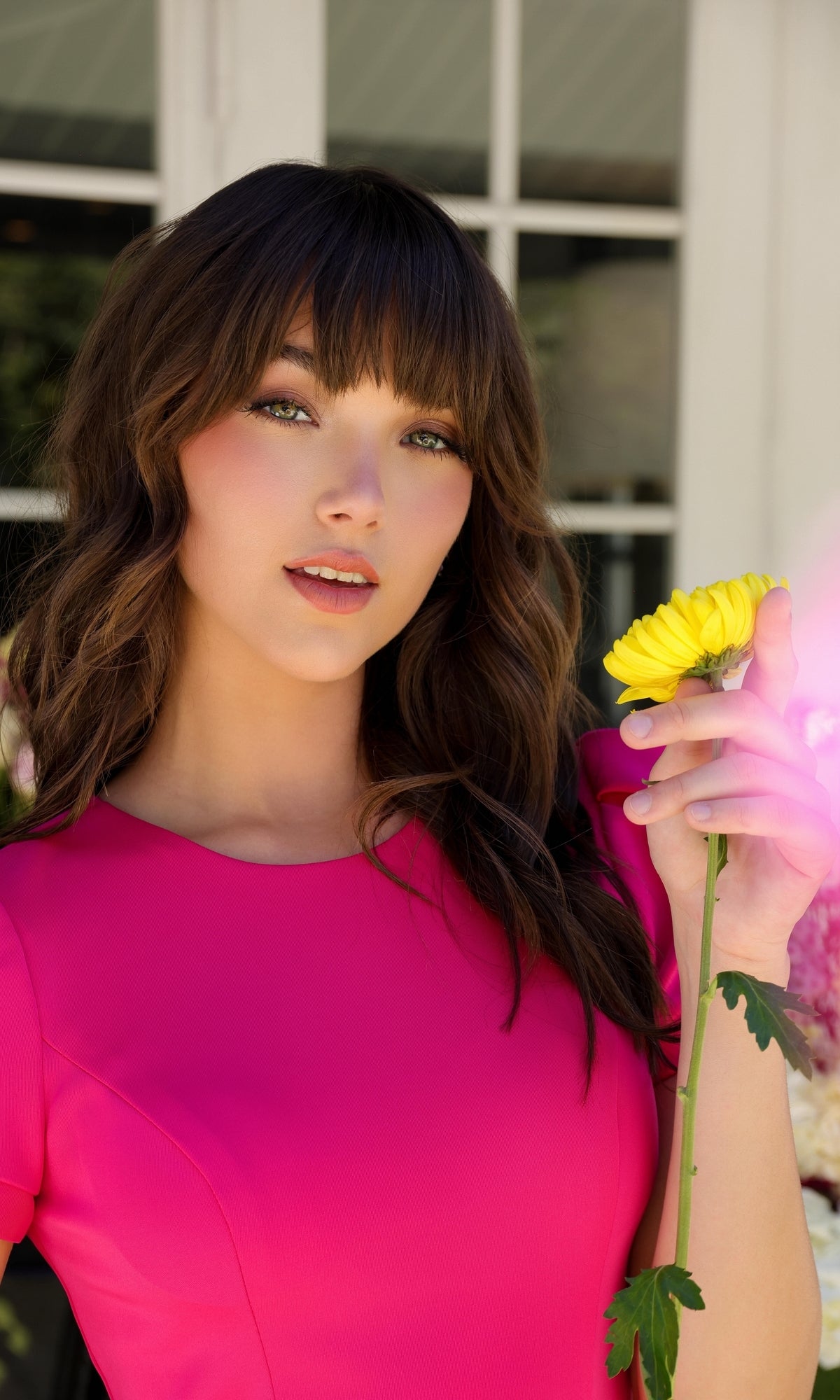 A woman with wavy brown hair in a bright pink Puff-Sleeve Homecoming Dress: Ava Presley 29801 holds a yellow flower, standing by a window with white frames.