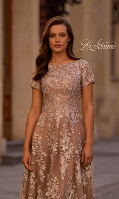A woman wears the Midi Mother of Bride Dress: La Femme 29830, a beige, short-sleeve midi dress with floral lace embroidery, standing outdoors and facing the camera.