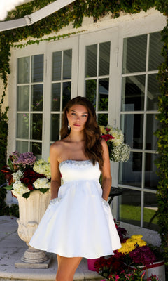 A woman wearing the Strapless Short Hoco Dress: Ava Presley 29837 stands by large windows, surrounded by potted flowers and lush greenery.