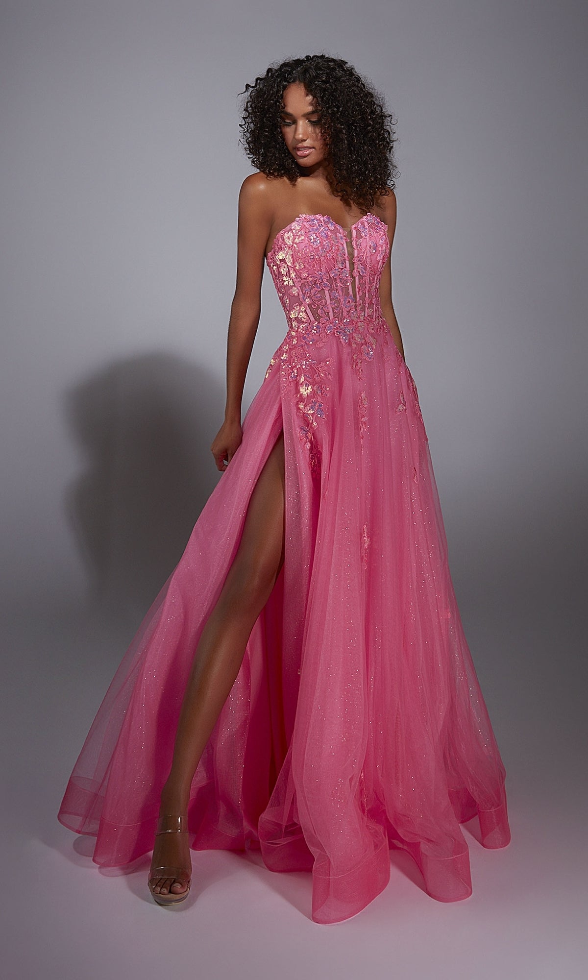 A woman poses against a plain background wearing the Aerie Couture 298 Glitter Tulle Prom Ball Gown, a strapless bright pink dress featuring floral lace embroidery, a sheer bodice, glitter tulle details, and a high leg slit.