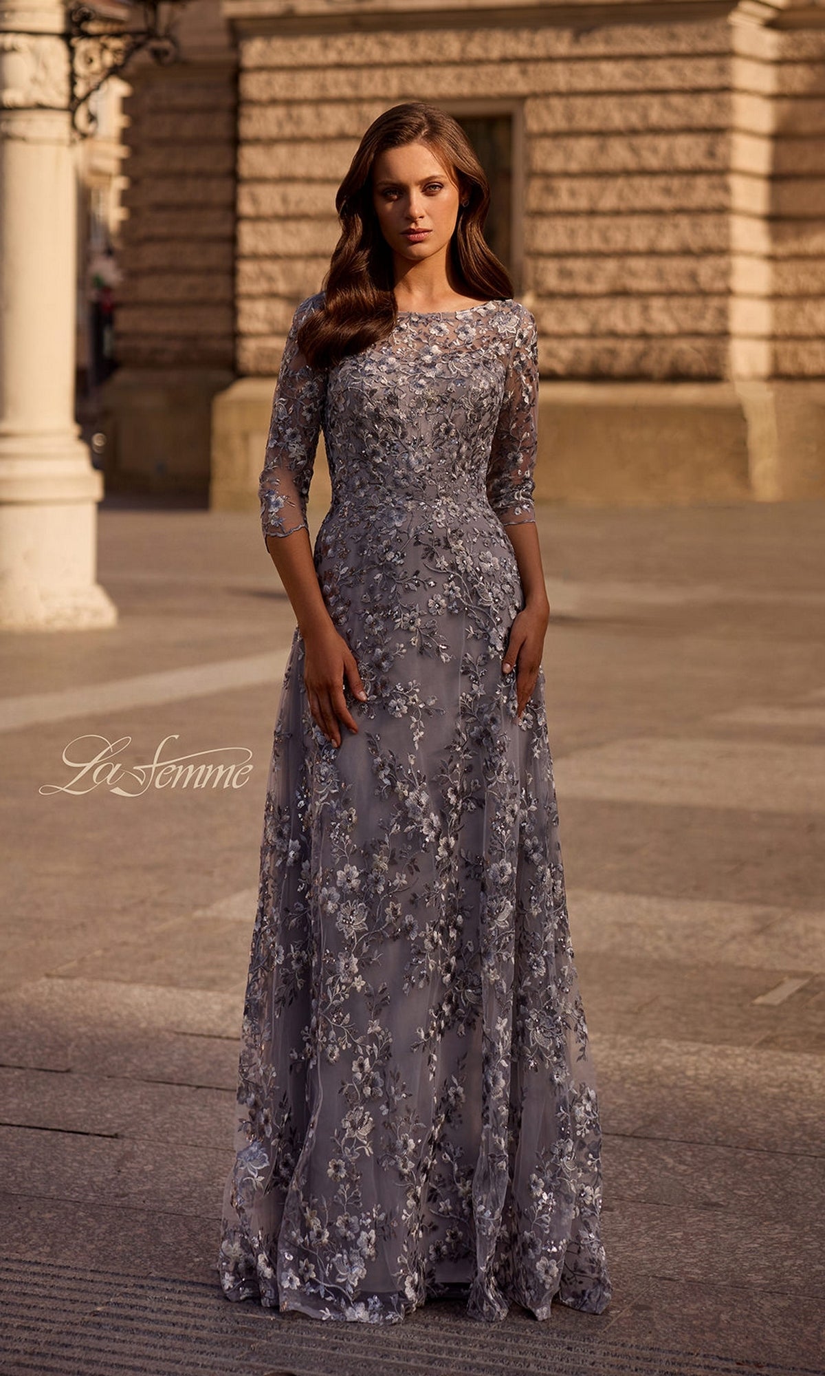 Woman stands outdoors in a silver Long Formal Dress: La Femme 29903 with metallic floral lace embroidery. Stone building and street lamp in the background.