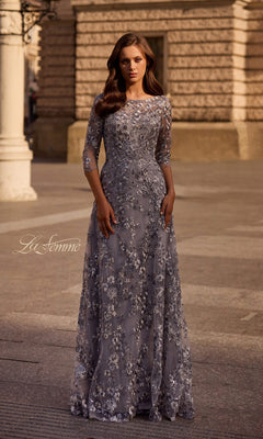Woman stands outdoors in a silver Long Formal Dress: La Femme 29903 with metallic floral lace embroidery. Stone building and street lamp in the background.