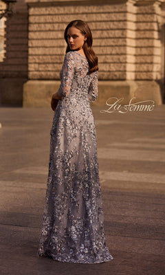 A woman in the Long Formal Dress: La Femme 29903, silver with metallic floral lace, stands outside facing away, with a stone building behind her.
