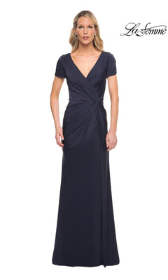 A woman wears a navy Long Formal Dress: La Femme 29926 featuring a V-neckline, short sleeves, and a draped, twist-knot waist.