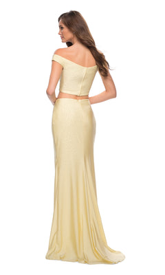 A woman stands with her back turned, wearing the La Femme 29951: an off-the-shoulder, light yellow two-piece prom dress with a fitted top and a long, flowing skirt.