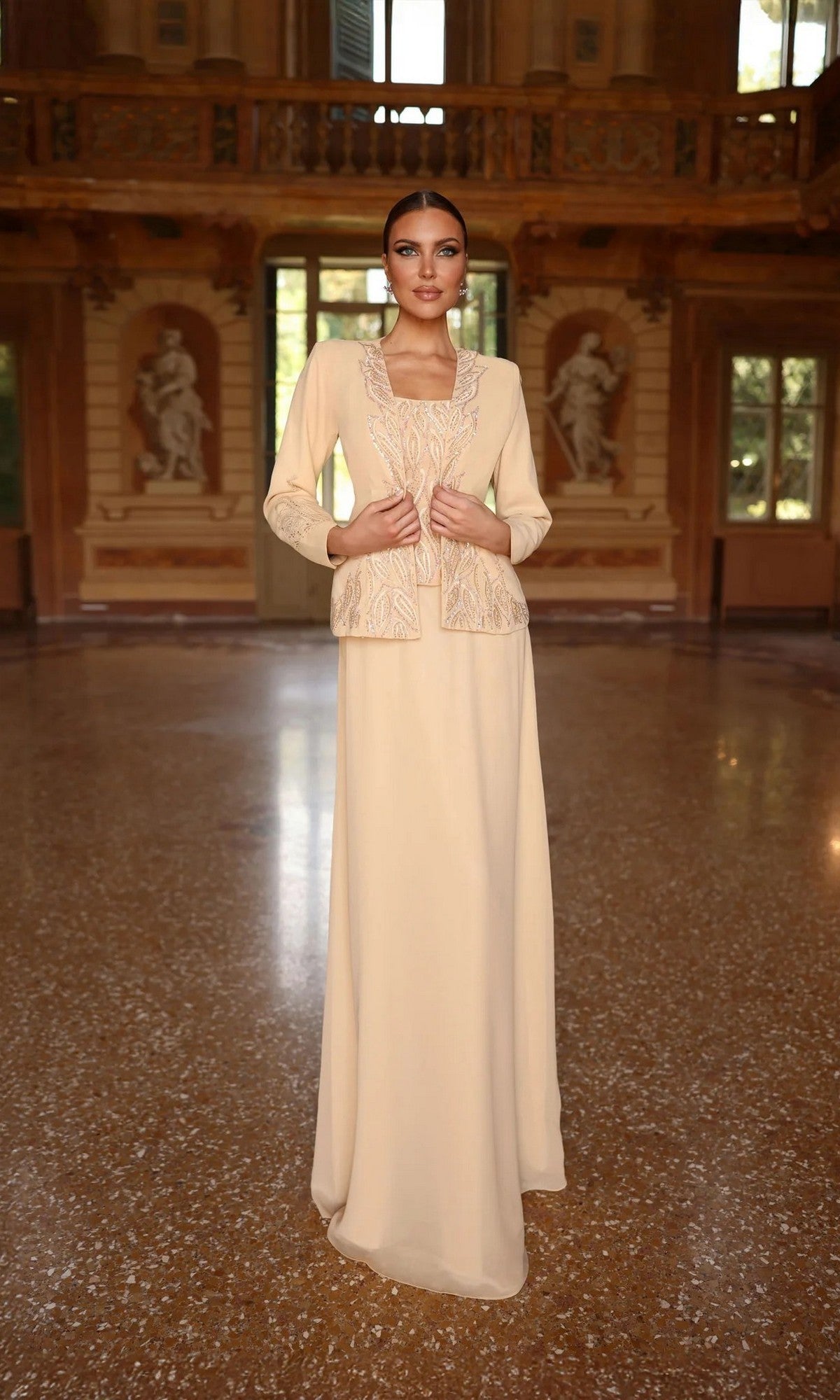 A woman stands in a grand hall wearing the Alyce 29953 long formal dress—a cream gown with a beaded top and ornate jacket—an elegant choice for the mother of the bride, framed by statues and large windows in the background.