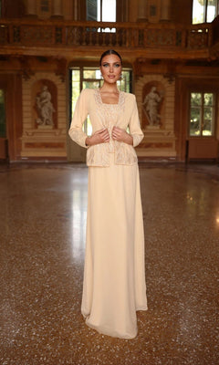 A woman stands in a grand hall wearing the Alyce 29953 long formal dress—a cream gown with a beaded top and ornate jacket—an elegant choice for the mother of the bride, framed by statues and large windows in the background.