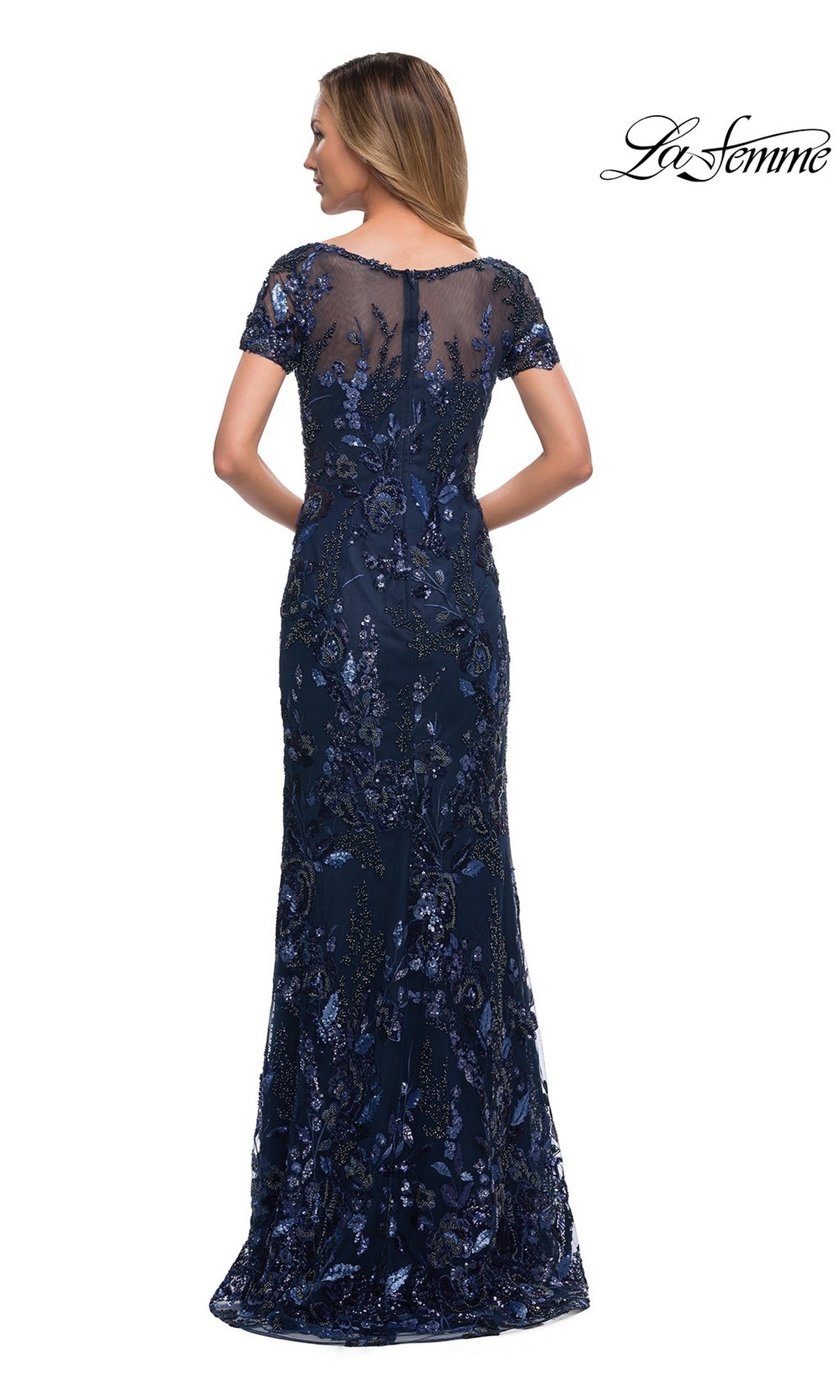 Woman in Long Formal Dress: La Femme 29961, navy blue sheath gown with sheer back and sequin floral embroidery, standing with her back to the camera.
