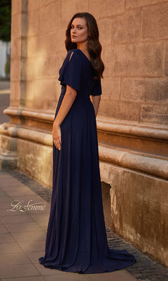 A woman in the Split-Sleeve Long Formal Dress: La Femme 30009 stands on a sidewalk by a stone wall, looking over her shoulder.
