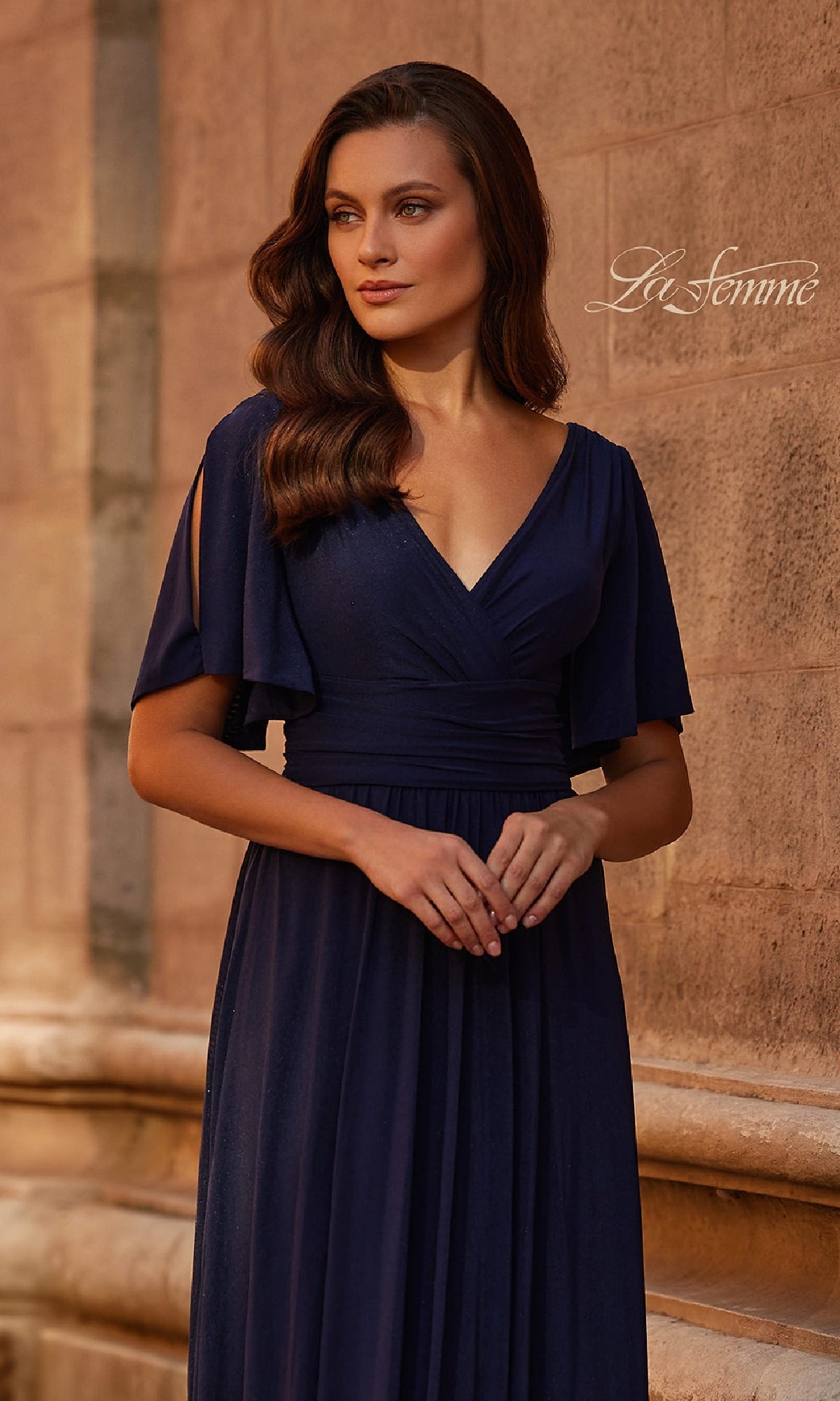 A woman wearing the Split-Sleeve Long Formal Dress: La Femme 30009 in navy blue stands against a stone wall, gazing to the side. 