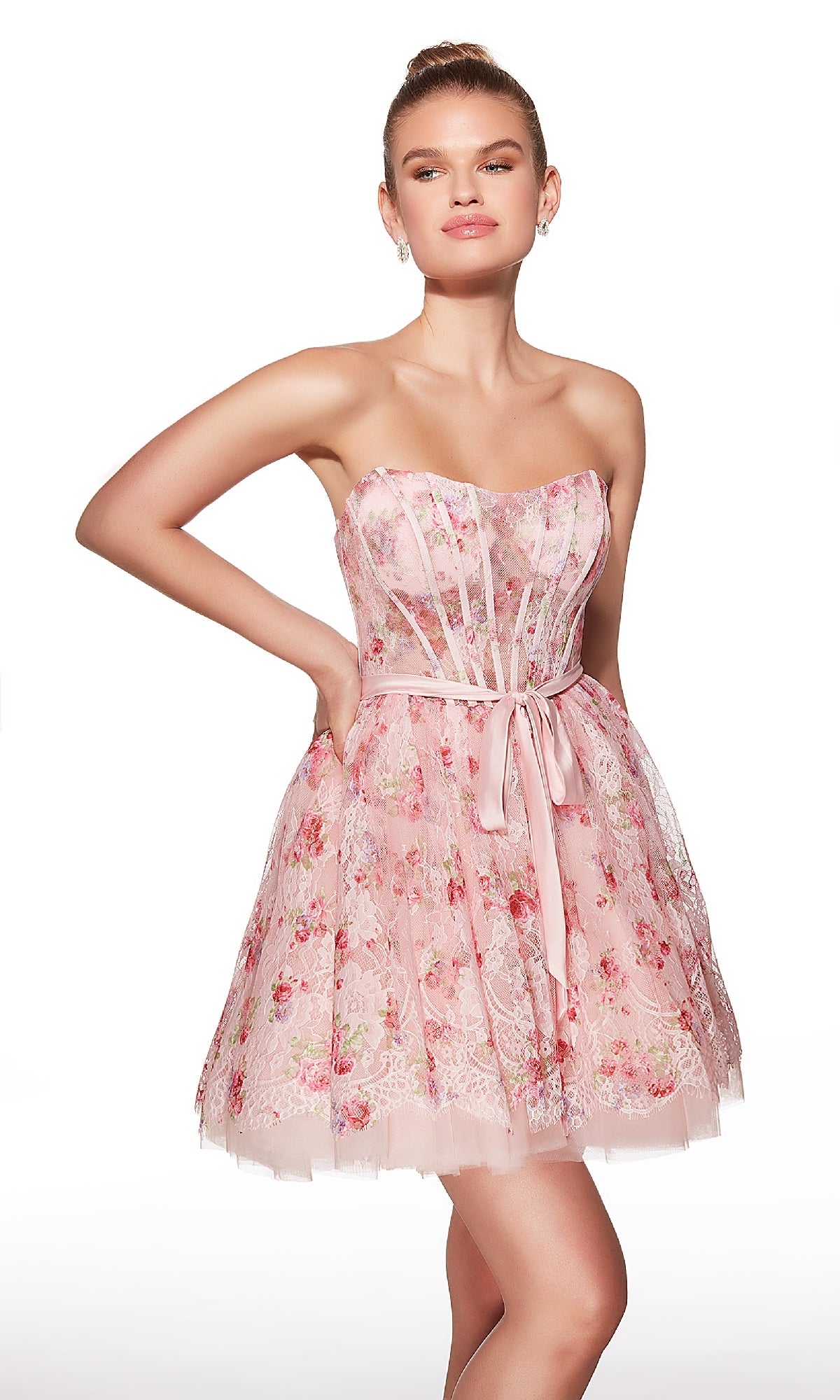 A woman wears the Alyce 30019 short homecoming dress—a pink floral fit-and-flare style with a sheer corset bodice and ribbon belt—while standing against a white background.