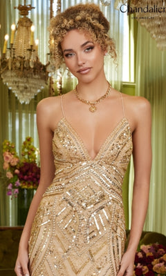 A woman wears the Long Prom Dress: Chandalier 30023, a gold sequin gown with thin straps, standing indoors near chandeliers and floral arrangements as the shimmering dress catches the light.