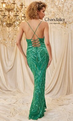 A woman in the Chandalier 30023 long prom dress with green sequins, a lace-up open back, and crisscross straps stands before chandeliers and draped fabric.