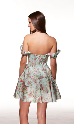 A woman faces away in the Alyce 30037 short homecoming dress—light blue with a pink floral pattern, off-shoulder neckline, fitted bodice, and flared skirt—against a plain white background.