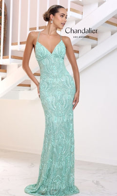 A woman stands indoors by a white staircase, wearing the Long Prom Dress 30047 by Chandalier—a fitted, light blue-green sequin formal gown with spaghetti straps.
