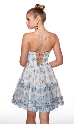 A woman stands facing away against a white background in the Lace-Up Short Print A-Line Hoco Dress: Alyce 30057, featuring blue and white florals, a crisscross back, and ruffled hem.