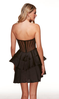 A woman with wavy blond hair and heart-shaped earrings stands with her back to the camera, wearing the Alyce 30084 Strapless Ruffled Short Hoco Dress featuring a sheer corset bodice and layered ruffled skirt—perfect for any party.