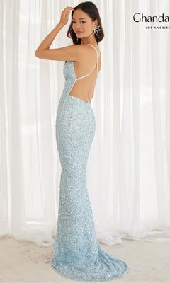 A woman stands in front of white sheer curtains wearing the Backless Long Sequin Prom Dress: Chandalier 30086, a light blue, strappy, open-back gown adorned with sequins.