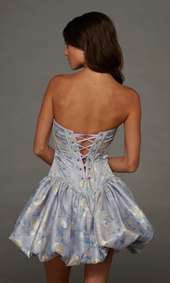 A woman wears the Alyce 30091 Short Homecoming Dress—a light blue metallic jacquard dress adorned with delicate floral patterns, featuring a strapless neckline, lace-up corset back, and playful bubble skirt—as she stands against a plain gray background.