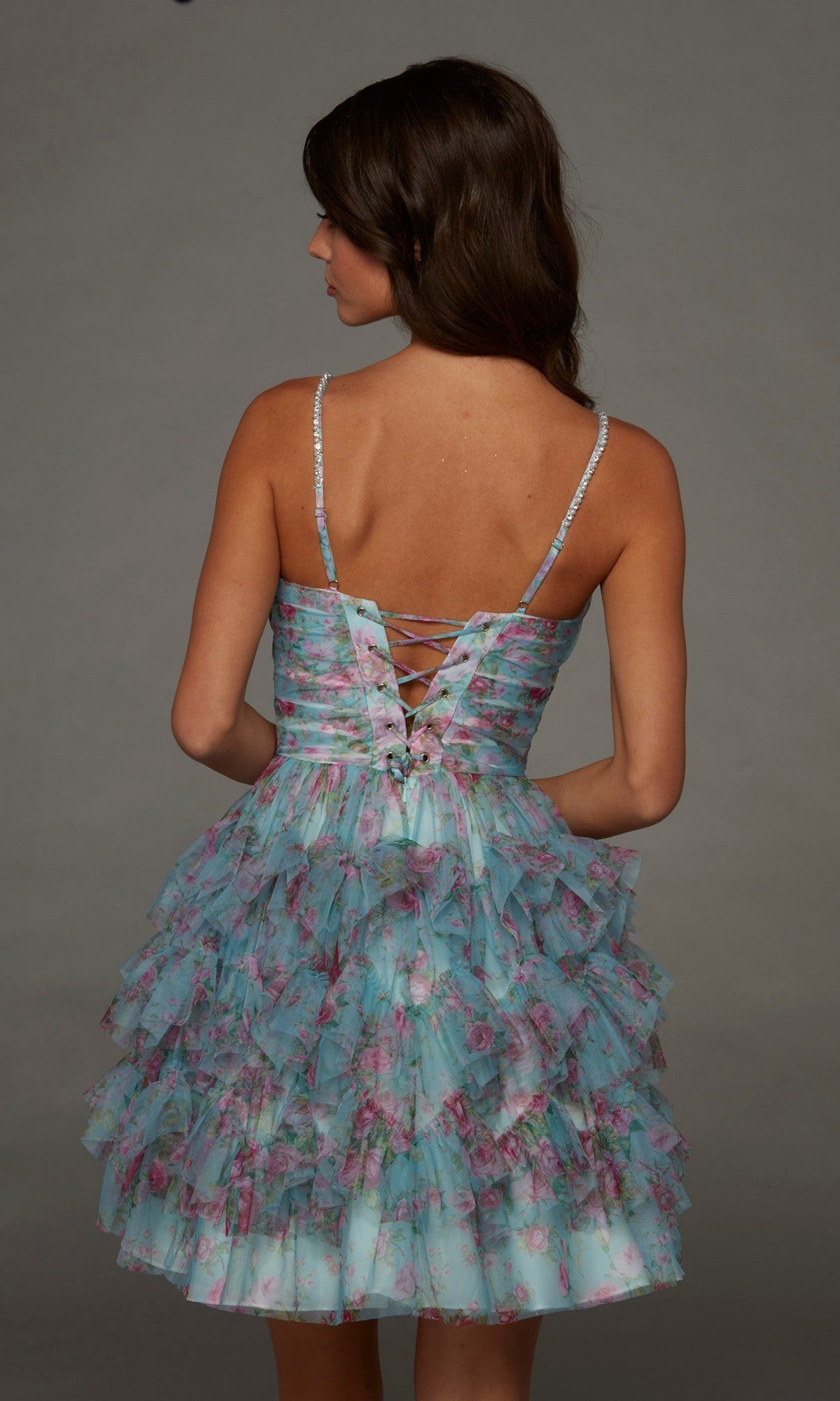 Facing away, the woman stands against a plain grey background wearing the Alyce 30092 short homecoming dress—a sleeveless blue floral design featuring ruffled layers and a lace-up back.