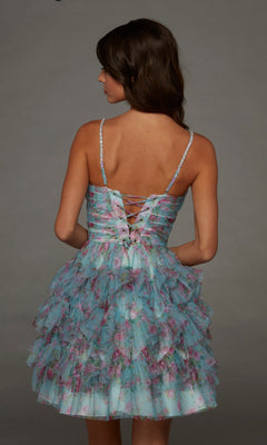 Facing away, the woman stands against a plain grey background wearing the Alyce 30092 short homecoming dress—a sleeveless blue floral design featuring ruffled layers and a lace-up back.