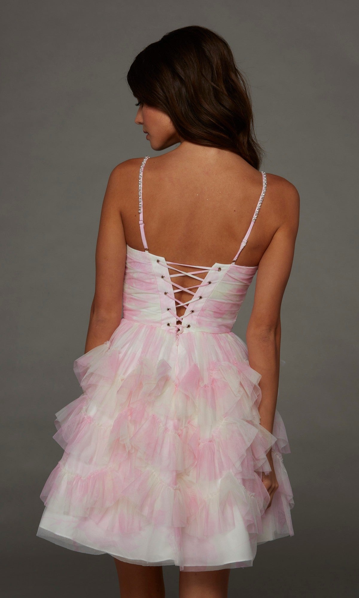 A woman is facing away, wearing the Alyce 30092 short homecoming dress featuring pink and white ruffles, lace-up back detailing, and spaghetti straps, set against a plain gray background.