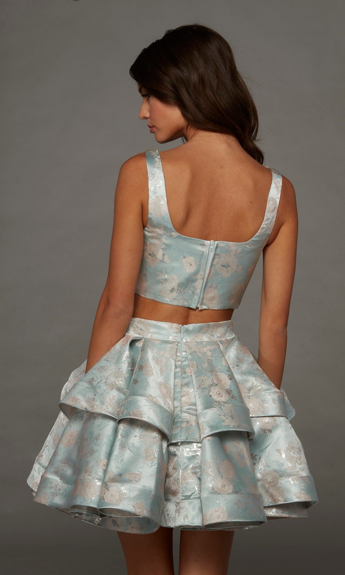 A woman faces away, modeling the Alyce 30095 short homecoming dress—a sleeveless, light blue floral two-piece set featuring a matching ruffled A-line skirt and a visible back zipper—against a plain gray background.