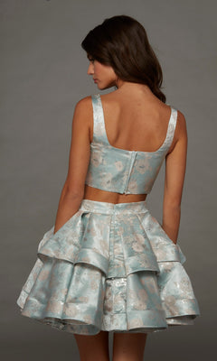 A woman faces away, modeling the Alyce 30095 short homecoming dress—a sleeveless, light blue floral two-piece set featuring a matching ruffled A-line skirt and a visible back zipper—against a plain gray background.
