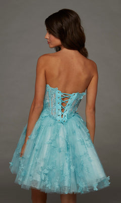 A woman with brown hair wears the Short Homecoming Dress: Alyce 30097—a light blue, strapless dress with a corset-back and delicate lace and floral details—while standing against a plain gray background.