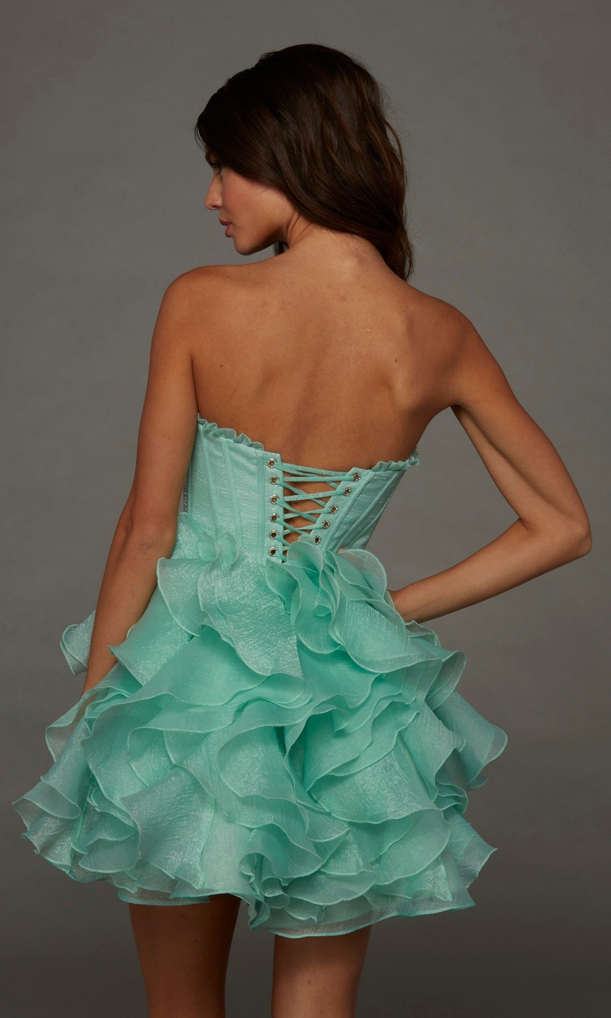 A woman is seen from behind, standing against a gray background, wearing the Alyce 30098 short homecoming dress—a strapless mint green A-line design featuring ruffled details and a lace-up back.