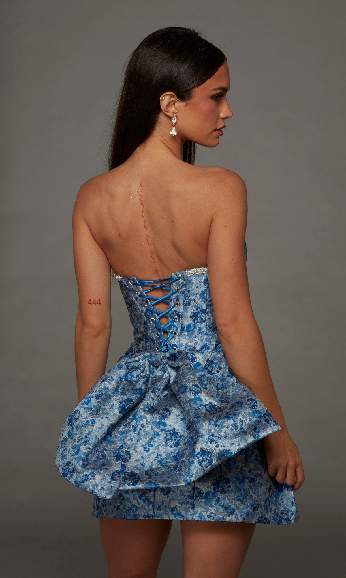 A woman stands turned away from the camera against a plain gray background, modeling the Alyce 30099 short homecoming dress—a blue floral strapless jacquard design featuring a lace-up back.