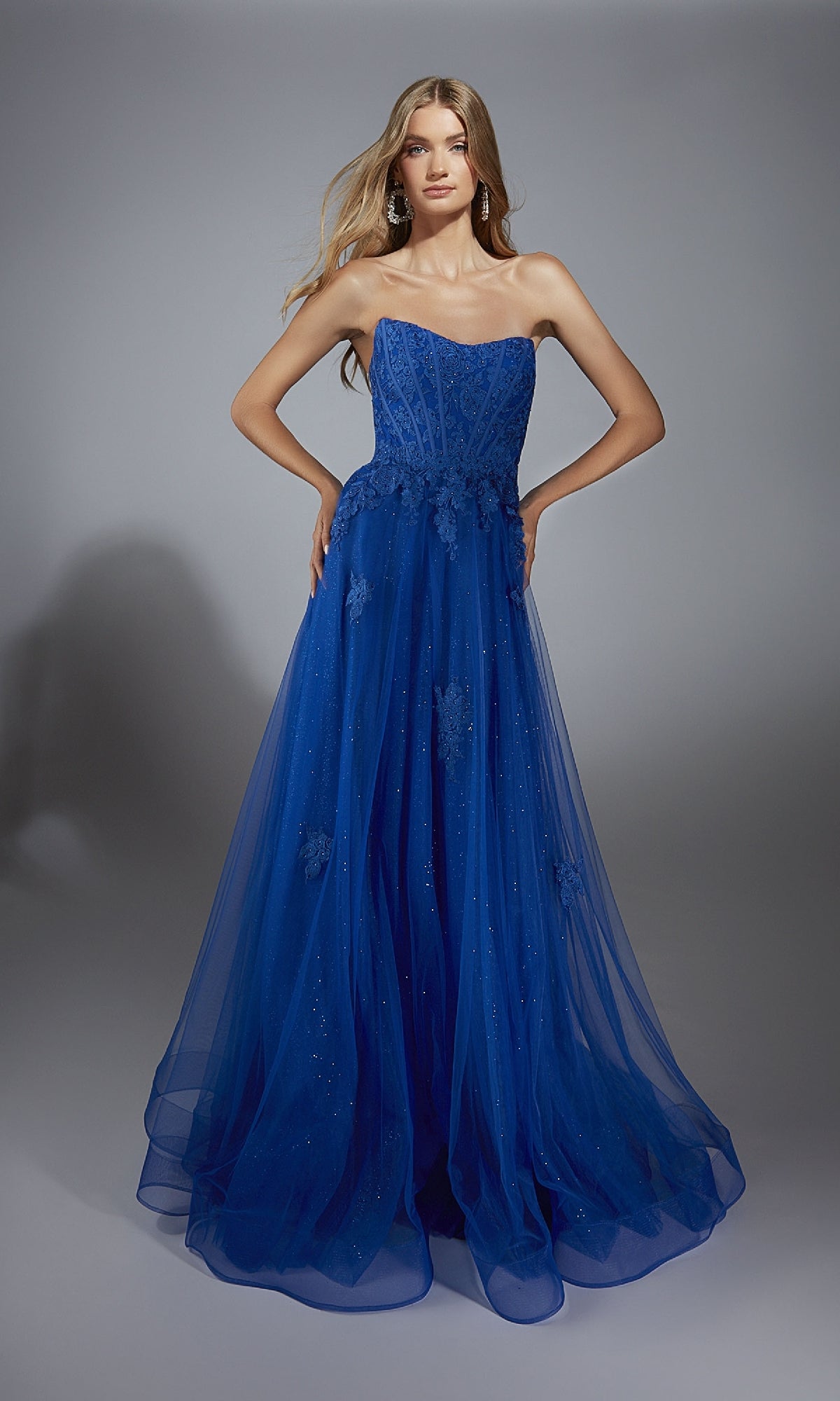A woman wearing the Aerie Couture 300 Long Prom Dress, featuring a strapless corset bodice, royal blue glitter tulle, floral appliqués, and a floor-length skirt, stands before a plain gray background.
