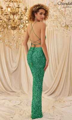 A woman wears the Backless Long Sequin Prom Dress: Chandalier 30101—a sparkly green gown with a strappy open back—standing before chandeliers and draped curtains.