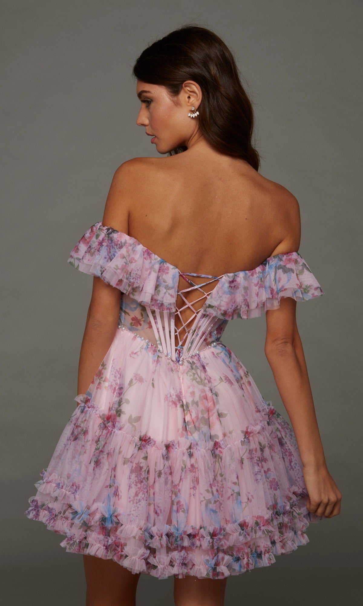 A woman wearing the Alyce 30103 short homecoming dress, featuring an off-shoulder design, pastel pink floral print, ruffled layers, and a lace-up back detail, stands against a plain background.