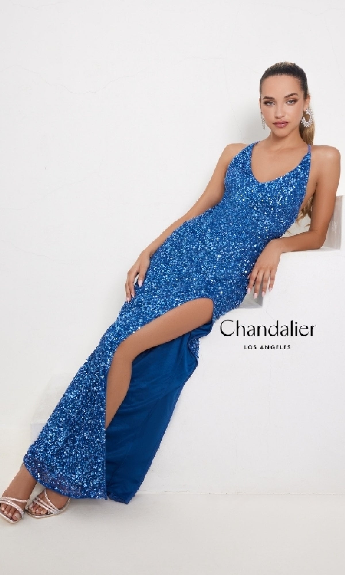 A woman sits against a white background in the Long Prom Dress 30103 by Chandalier, featuring a fitted sequin design, V-neck, and thigh-high slit. The 