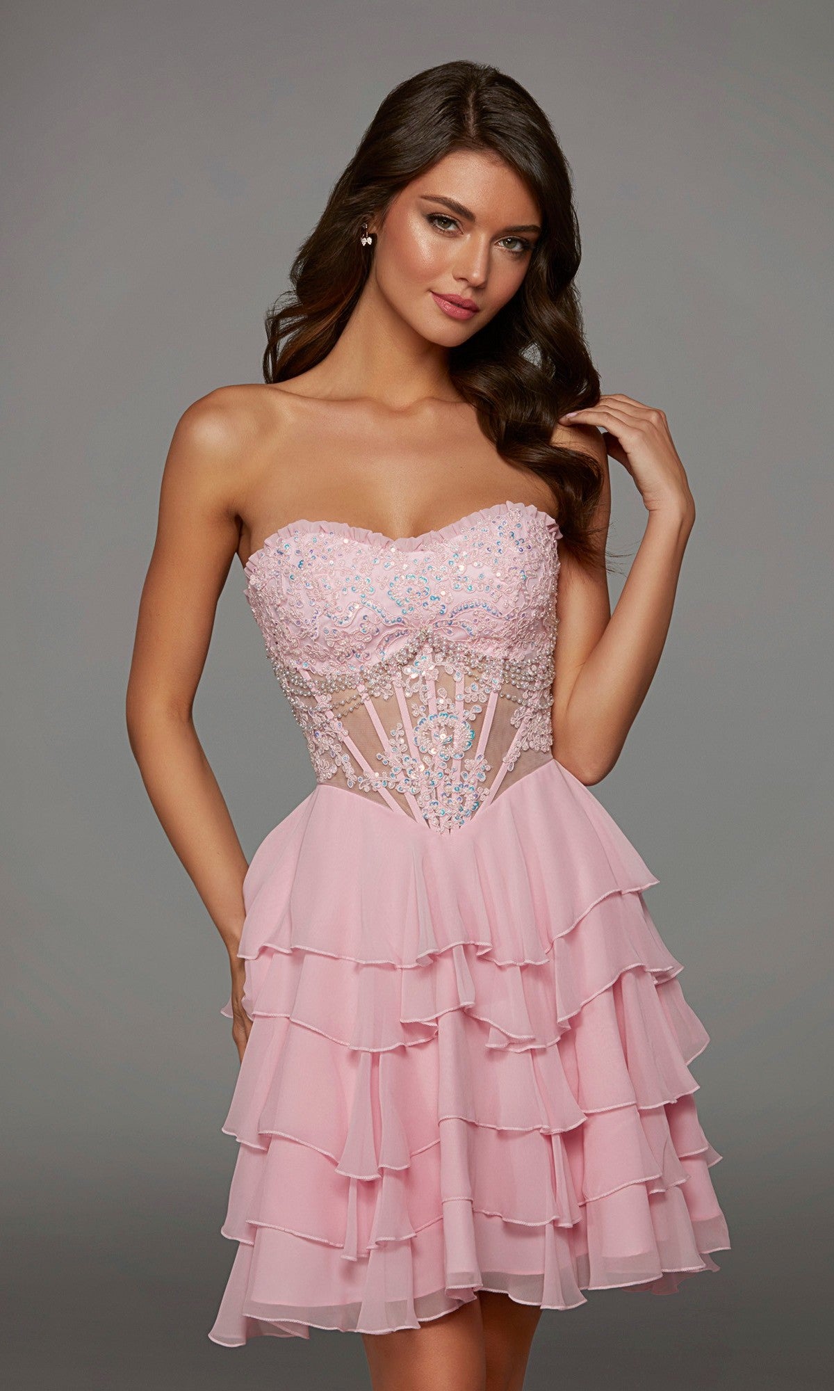 A woman wearing the Alyce 30104 short homecoming dress in pink, featuring a strapless beaded bodice, sheer waist, and tiered ruffle skirt, stands against a plain gray background.