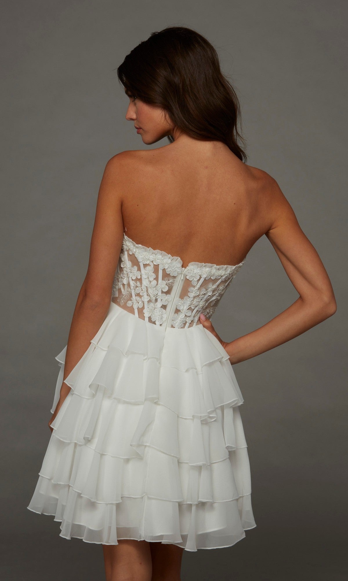 Wearing the Alyce 30109 short homecoming dress, a woman poses with her back to the camera against a plain gray background, showcasing the strapless fit-and-flare design with a lace bodice and tiered ruffle skirt.