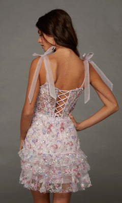 A woman poses against a plain gray background wearing the Alyce 30111 short homecoming dress, featuring a floral design with sequin-embellished details, a ruffled skirt, lace-up back, and ribbon shoulder straps.