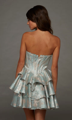 Woman in the Alyce 30112 short homecoming dress, featuring a strapless fit-and-flare silhouette with a metallic blue floral design and ruffled skirt, stands with her back to the camera against a gray background.