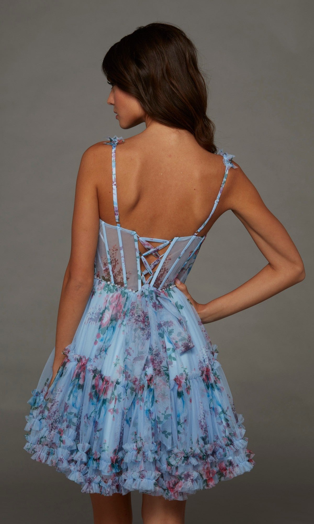 A woman stands with her back to the camera, wearing the Short Homecoming Dress: Alyce 30113, a light blue floral-printed dress featuring a lace-up corset bodice and ruffled skirt accents.