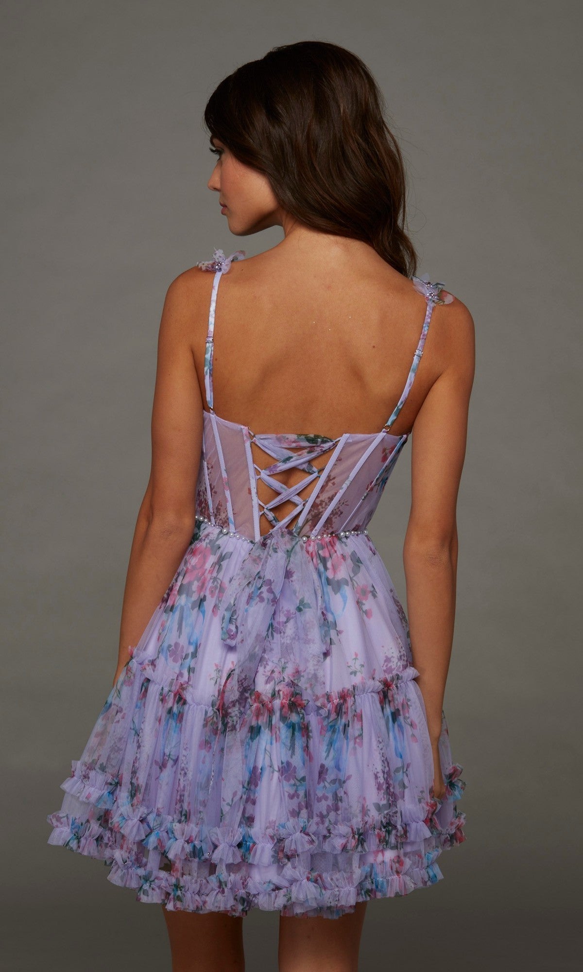 A woman stands facing away against a plain gray background, wearing the Alyce 30113 short homecoming dress—an elegant lavender floral-print style featuring ruffled layers and a lace-up corset bodice.