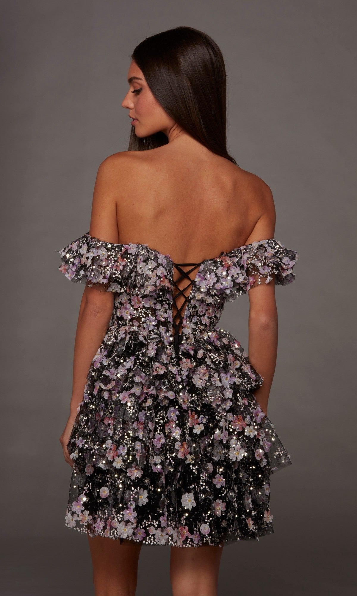 A woman faces away against a plain gray background, wearing the Alyce 30115 Short Homecoming Dress—an off-shoulder, ruffled, floral-patterned fit-and-flare party dress featuring a lace-up back and a layered skirt.